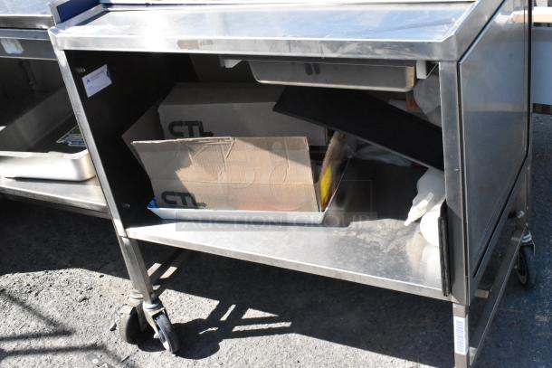 Stainless steel cart with under shelf on commercial casters, featuring visible cardboard box and tray, in used condition.