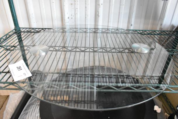 Round glass pane on metal shelf, clear, smooth surface, two circular metal weights on top, auction tag reading "261".