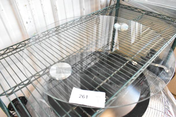 Round glass pane with smooth edges, in excellent condition, two metallic stands underneath, auction tag number 261 visible.