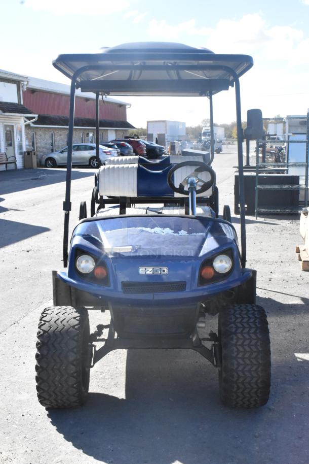 EZ Go blue and white 6-seat golf cart with speaker system, showing minor wear, featuring a sturdy roof and large tires.