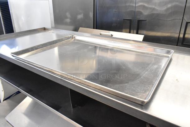 24 full-size metal baking pans on a stainless steel surface, in used condition, suitable for commercial baking.