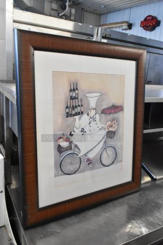Framed artwork depicting a chef on a bicycle with wine bottles and bread. Dark wooden frame in good condition.