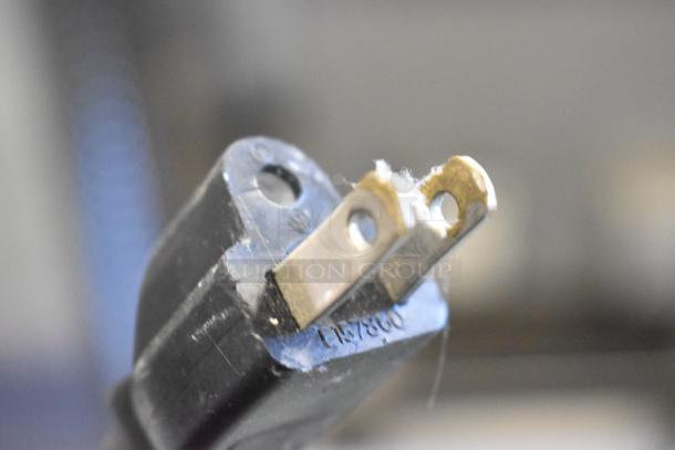 Close-up of a black electrical plug with visible prongs and markings, part of a Lowe metal commercial deli display case.