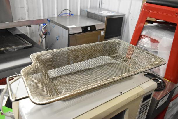 Metal mesh strainer basket resting on a microwave, surrounded by stainless steel kitchen equipment in a storage area.