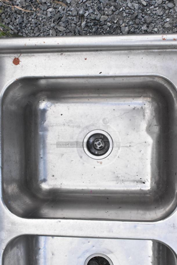 Stainless steel 3 bay sink without legs, visible scratching and rust spots, built-in drain, durable material.