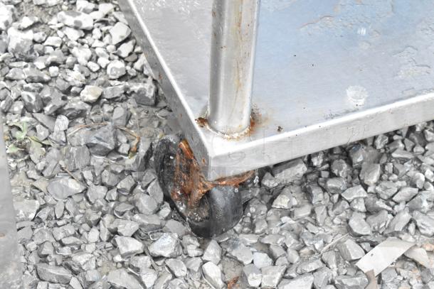 Stainless steel equipment stand on rusted commercial caster, showcasing under shelf. Condition suggests outdoor exposure.