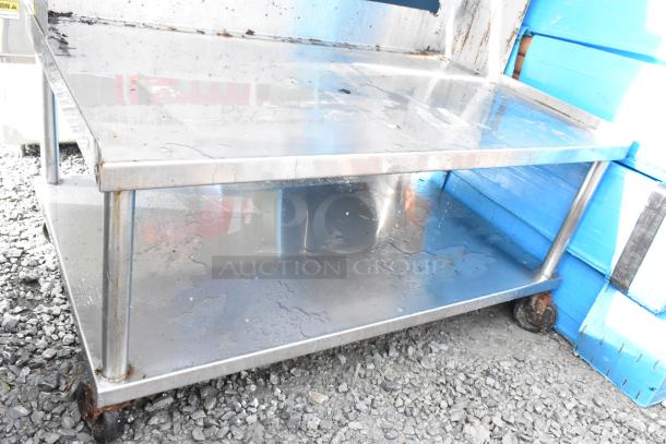 Stainless steel equipment stand with over and under shelves, on commercial casters, shows signs of wear and rust.