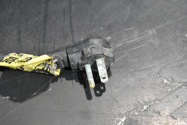 Frayed black electrical plug with yellow caution tag attached. Plug shows signs of wear and corrosion.