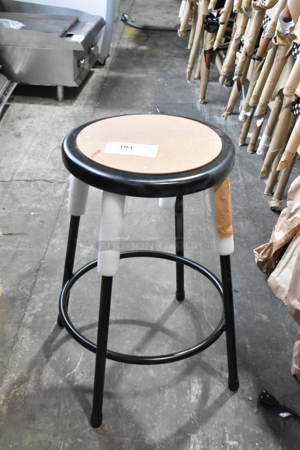 Brand new black metal stool, scratch and dent, with protective packaging; shows label "104" on the seat top.