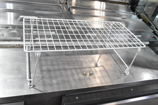 White metal wire rack with foldable legs on stainless steel surface. Labels in background. Good condition.