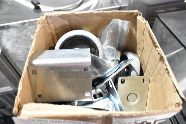 Lot of 4 brand new commercial casters in a cardboard box, showing minor scratches and dents, metal and rubber construction.