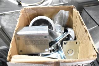 Lot of 4 brand new commercial casters in a cardboard box, showing minor scratches and dents, metal and rubber construction.