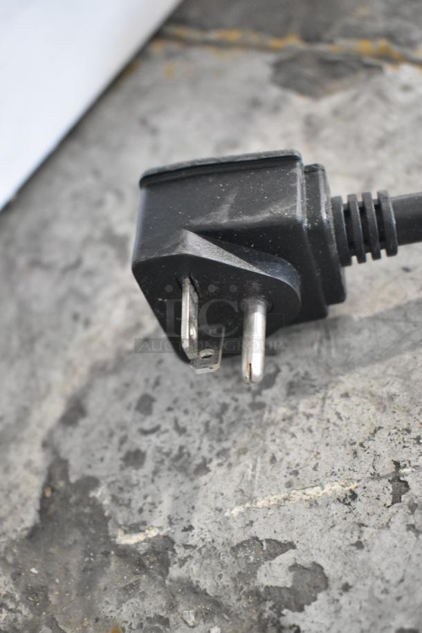 Close-up of a two-prong plug attached to the Menumaster RFS12TSW commercial microwave oven. Dark plastic housing, slight wear.