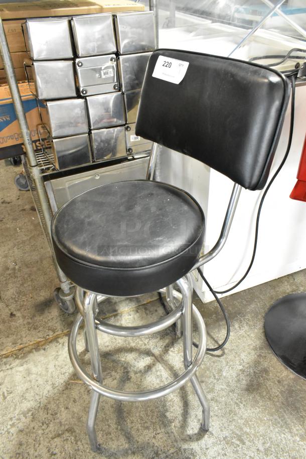 Black stool with backrest on metal frame, featuring a padded seat and chrome legs. Minor wear visible on upholstery.