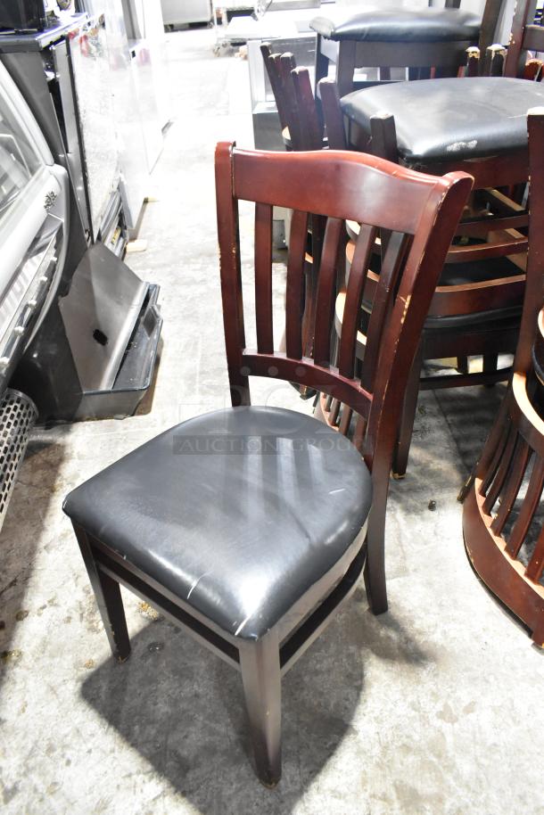 14 wooden dining chairs with black seat cushions, showing wear. Ideal for refurbishing or use. Auction lot of 14.
