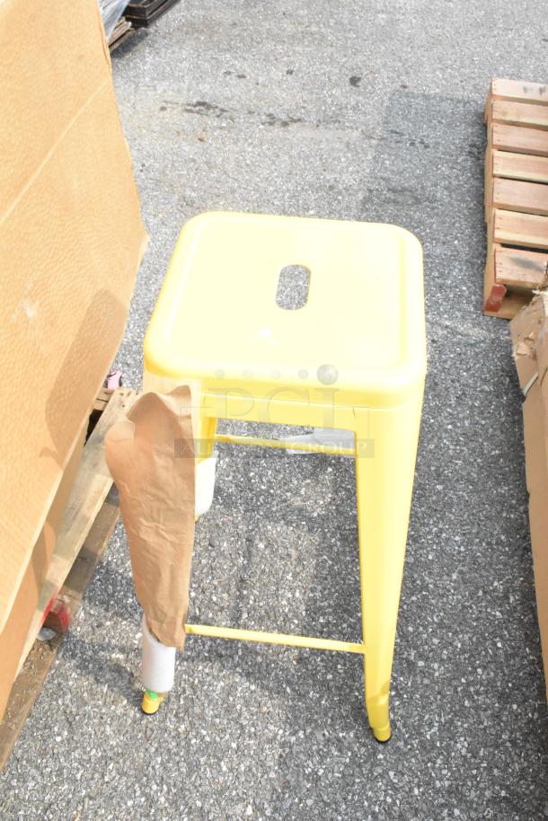 Yellow Tolix style stool, brand new, with square seat and footrests, shown on a pallet outdoors.