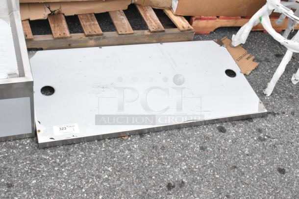 Brand new stainless steel panel with visible scratches and dents, resting on the ground, marked with label "323 1 QTY".