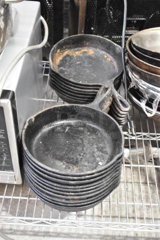 Stacks of 17 vintage cast iron skillets on metal shelving, showing wear and patina, ideal for restoration collectors.