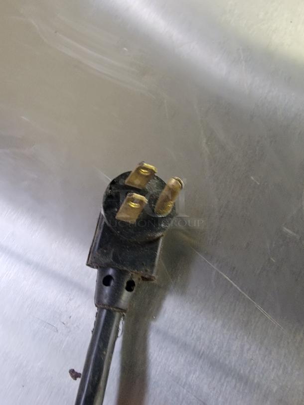 Close-up of a three-prong electrical plug attached to a cord, resting on a stainless steel surface.