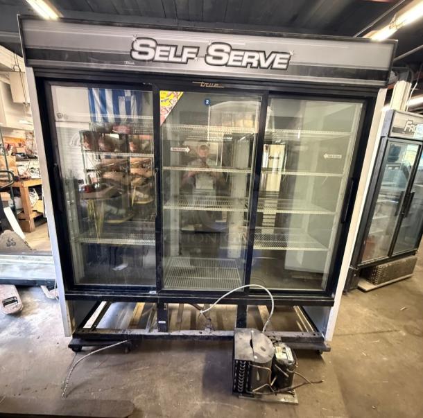 True GDM-69 three-glass sliding door commercial refrigerator. Features shelving, "Self Serve" label, visible wear.