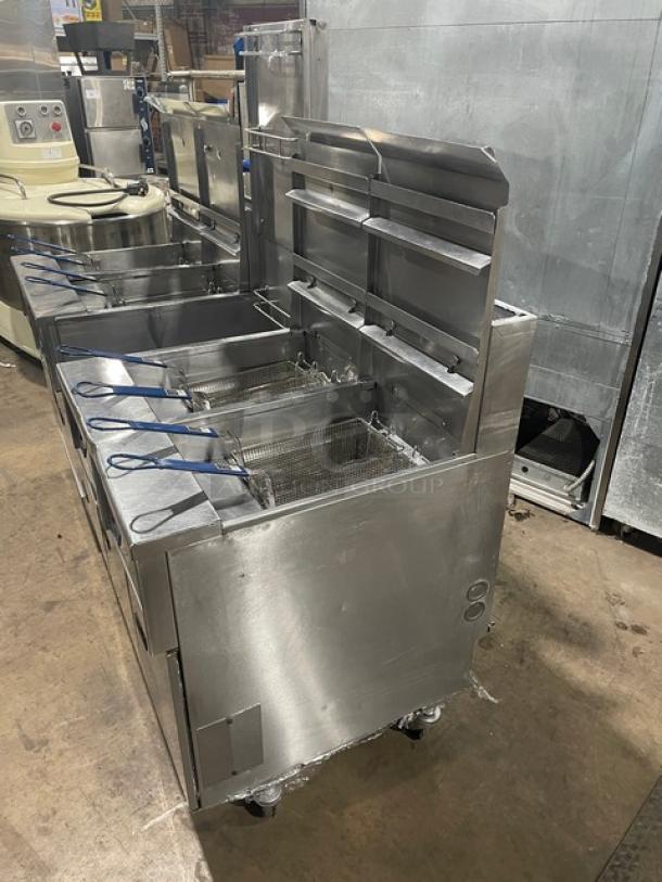 Pitco FGSGH50 stainless steel 4-bay fryer system, natural gas, 50 lb. capacity. Includes basket hangers, backsplash, oil filter, and blue gas hose. Mounted on casters.