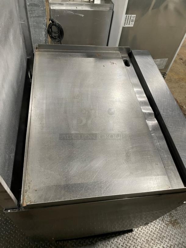 Migali commercial stainless steel natural gas flat griddle range/oven combo with backsplash and wired oven racks on casters.