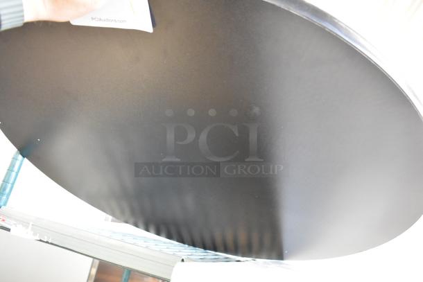 Brand new black round tabletop showing minor scratches and dents. Includes PCI Auctions tag.