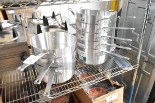 8 new Winco ASP-10 10 qt aluminum saucepans with solid handles, minor dents, on metal shelf. Tags indicate model.