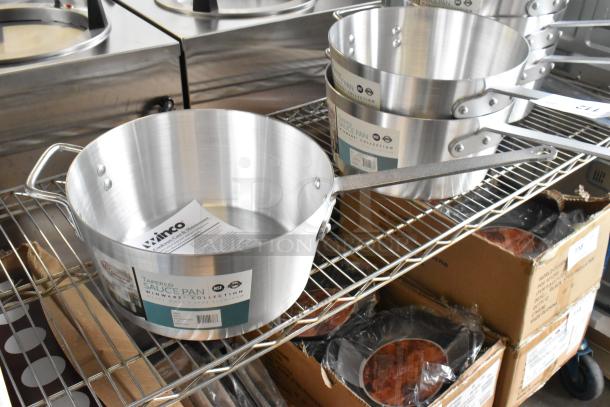 8 new Winco ASP-10 10 qt aluminum saucepans with solid metal handles, minor scratches, on shelf. Tags indicate durability.