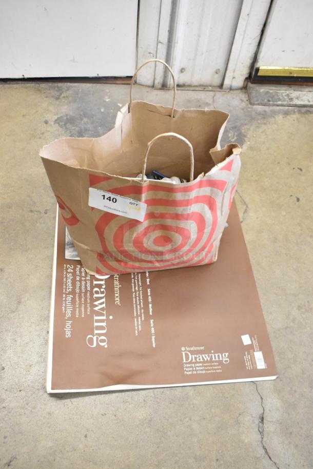Brown paper bag with handles, numbered tag "140," next to a Strathmore drawing pad, containing acrylic paints and brushes.