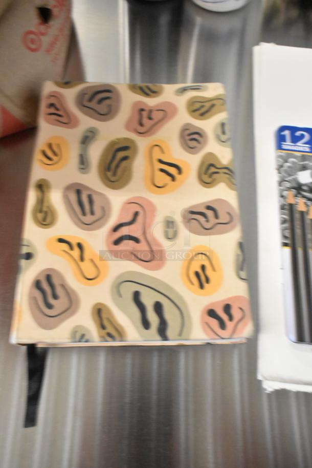 Lot of art supplies including a drawing pad with abstract cover, acrylic paints, and brushes. Ideal for artists.