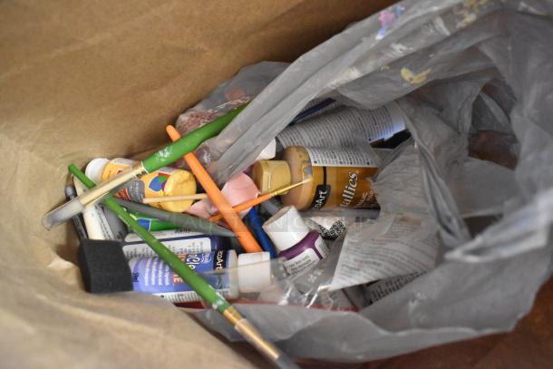 Lot of art supplies including various acrylic paints, brushes, and a drawing pad, loosely packed in a paper bag.