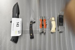 Five various pocket knives, including Schrade 1520T, CRKT R2301, Camillus 440, on a metal surface; auction tag stating "143 QTY 5" visible.