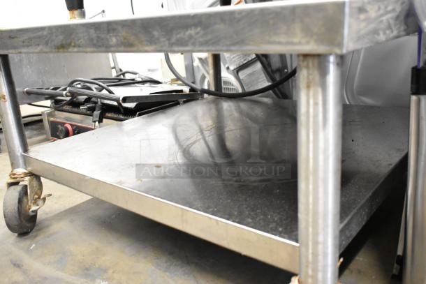 Stainless steel equipment stand with under shelf on commercial caster, showing signs of wear. Ideal for robust use.