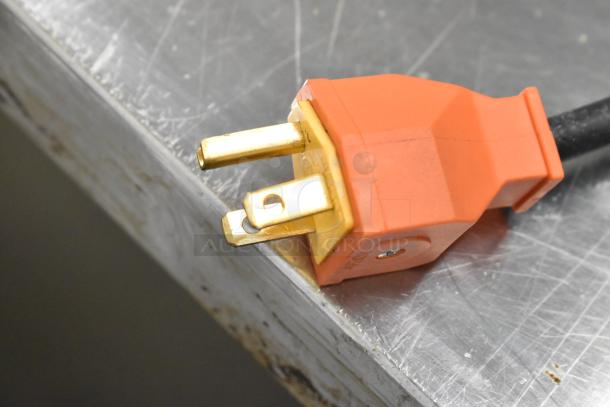Close-up of an orange electrical plug with three prongs, resting on a scratched metal surface.