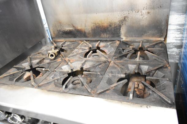 Stainless steel commercial gas range with 6 burners, oven, overshelf, backsplash, and casters. Shows signs of use.