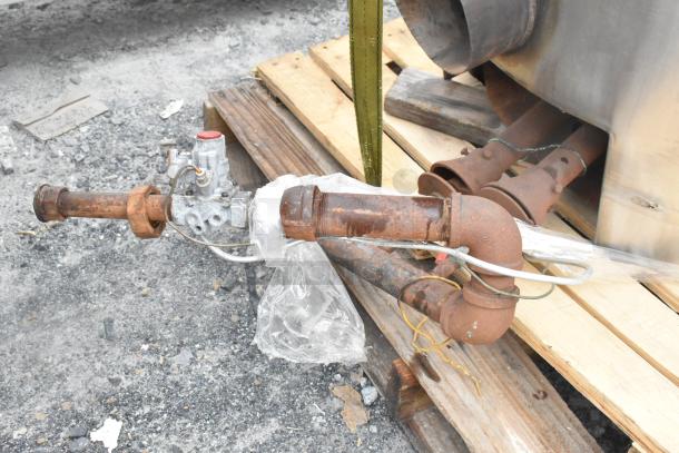 Stainless steel bagel kettle with attached rusted pipes, resting on wooden pallets, showing various fittings and hoses.