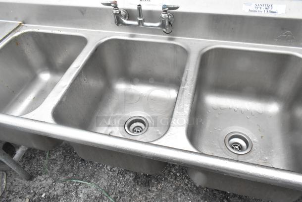 Stainless steel 3 bay sink with dual drain boards, handles, and visible faucet; clean, lightly worn condition.