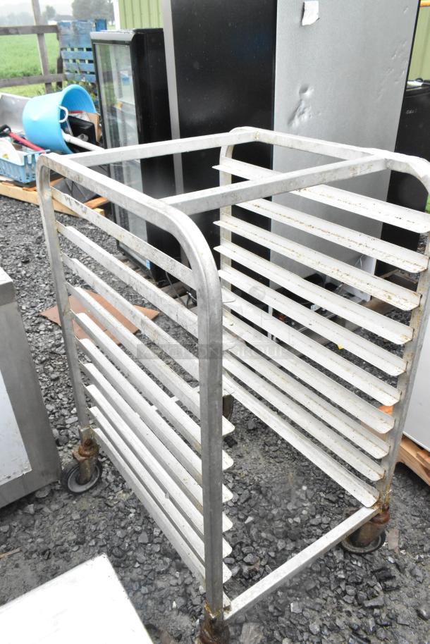Metal commercial transport pan rack on casters, silver, shows signs of wear, multiple shelves, outdoor setting.