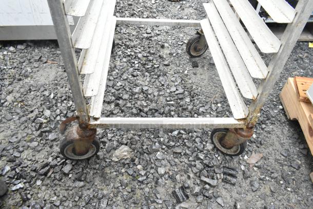 Metal commercial transport pan rack with multiple shelves on commercial casters, showing rust and wear, outdoor setting.