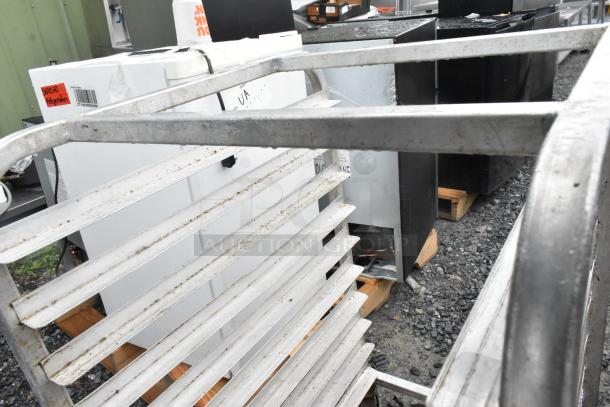Metal commercial transport pan rack on casters, featuring multiple shelves. Signs of wear and outdoor storage visible.