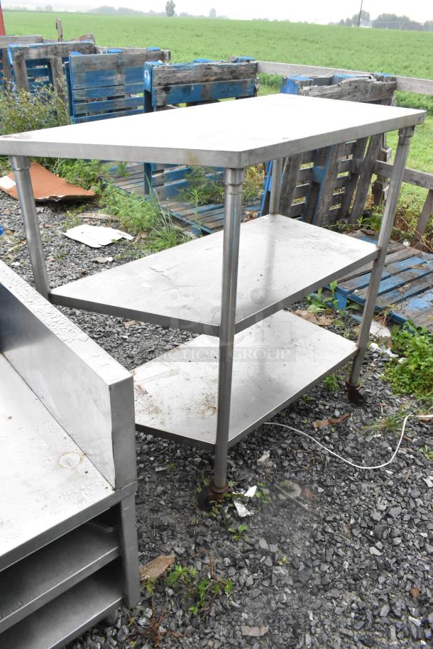 Stainless steel 3-tier shelving unit on commercial casters, outdoor setting. Some wear visible.