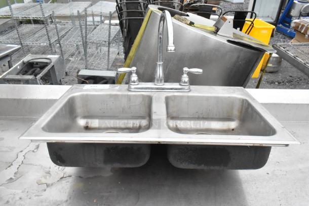Metal 2 bay sink with dual handles and high-arc faucet, featuring slight wear; suitable for commercial use.