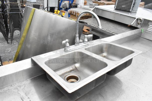 Dual-basin metal sink with high-arc faucet and dual handles. Slight wear visible. Ideal for commercial use. Marked "273".