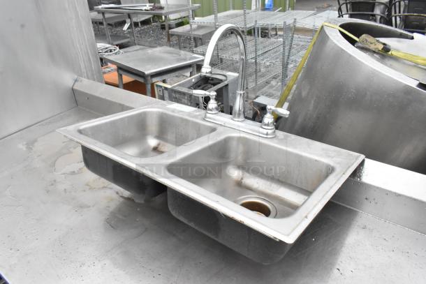 Metal 2 bay sink with faucet and handles, stainless steel, used condition, signs of wear, suitable for commercial use.
