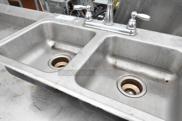 Metal 2 bay sink with faucet and handles, showing signs of wear. Includes attached label with item number.