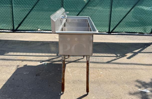 Stainless steel triple sink with three basins and a backsplash, mounted on rusted metal legs. Appears used with visible wear.
