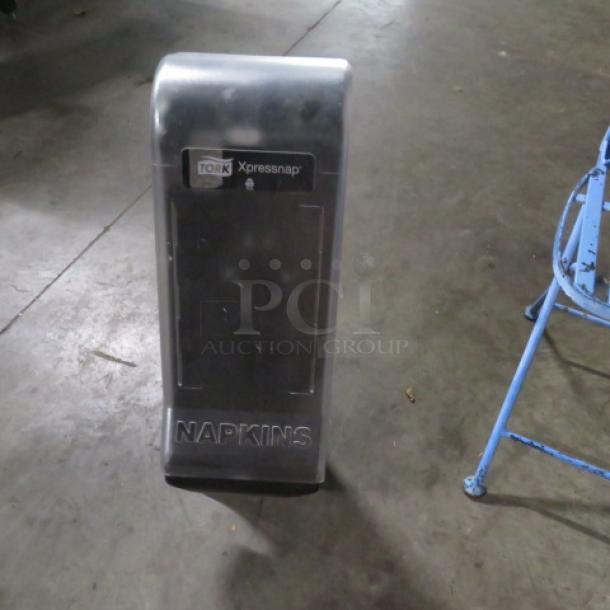 Tork Xpressnap napkin dispenser with visible branding and "NAPKINS" label, in used condition.