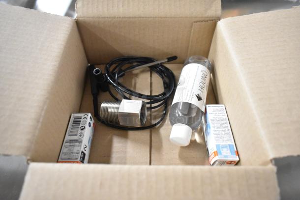A cardboard box containing various small items, including a coiled wire, metal connector, a bottle labeled "HOUNÖ," and two small boxed components.