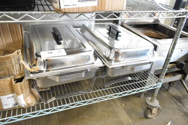 Two brand new stainless steel chafing dishes, minor scratches and dents, with black handles, placed on a metal rack.